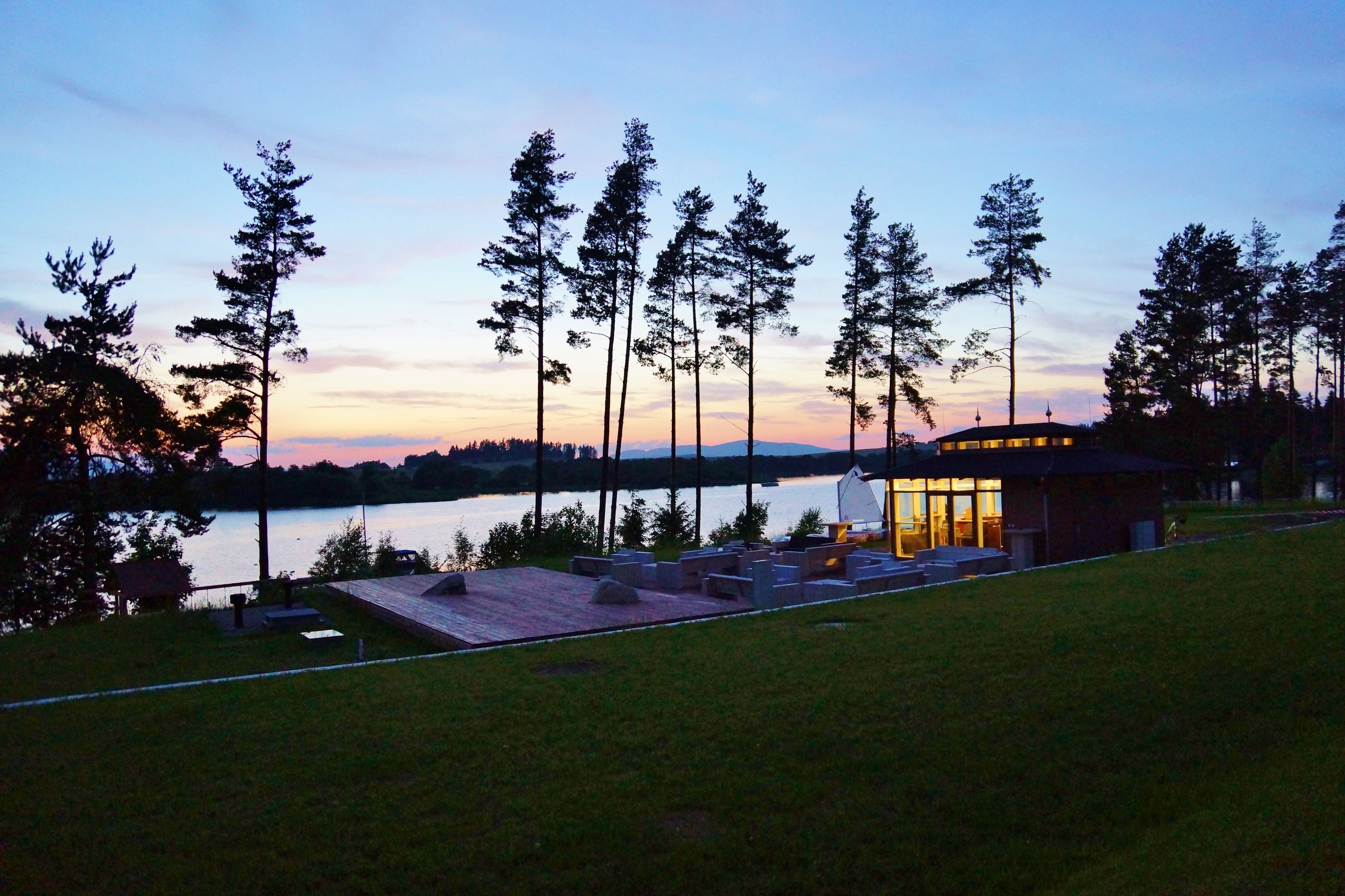 A lakeside property with a wooden deck, a small building with glass walls, and tall trees.