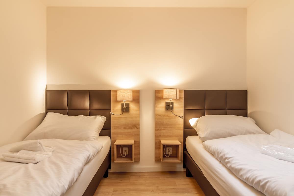A hotel room with two beds, wooden headboards, bedside tables, and wall-mounted lamps.
