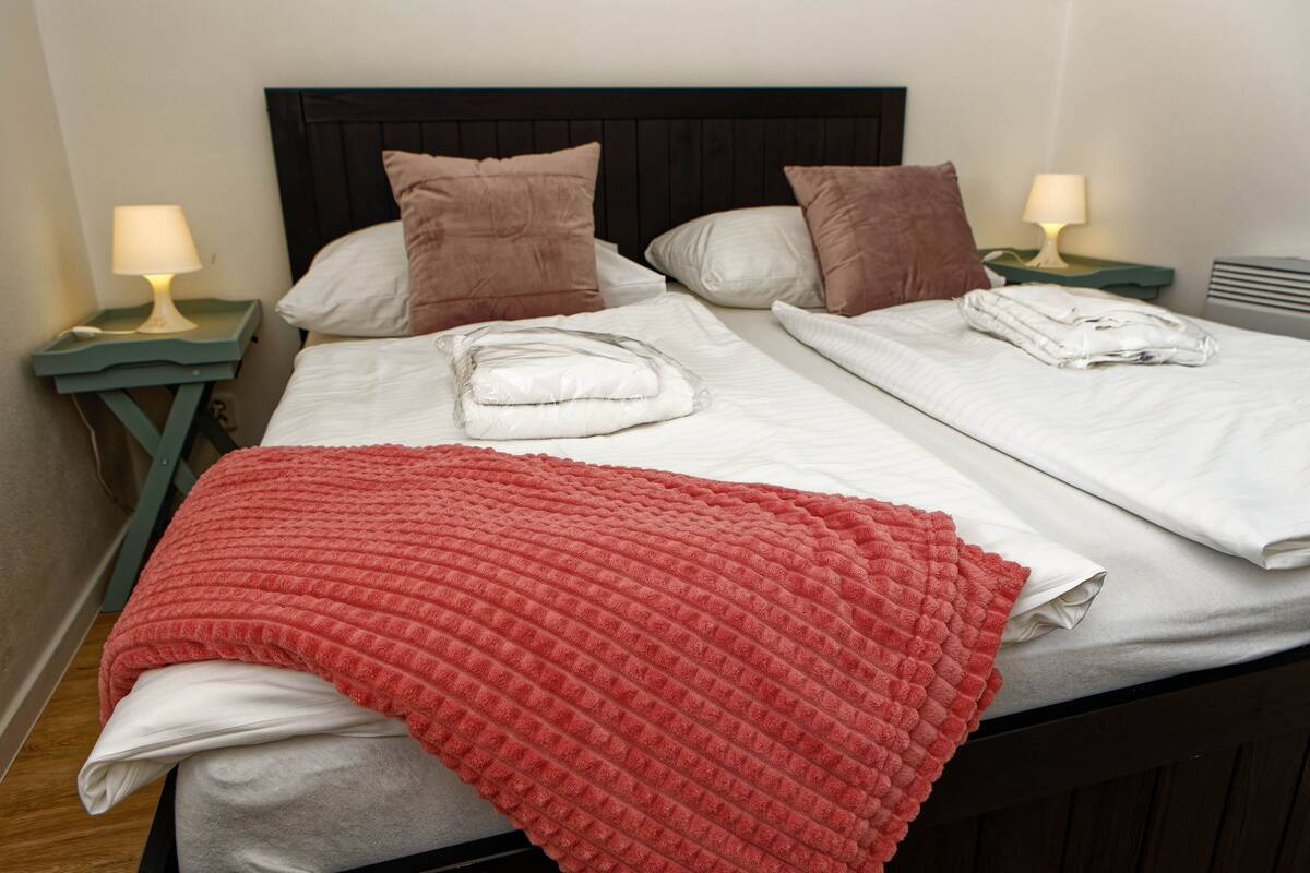 A neatly made bed with white linens, a red blanket, and two pillows. There are two bedside tables with lamps.