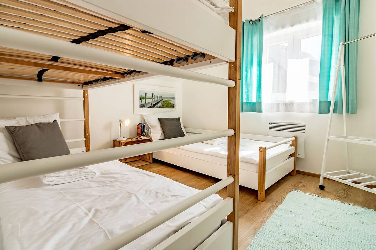 A dormitory room with bunk beds, a bedside table with a lamp, a window with curtains, and a radiator.