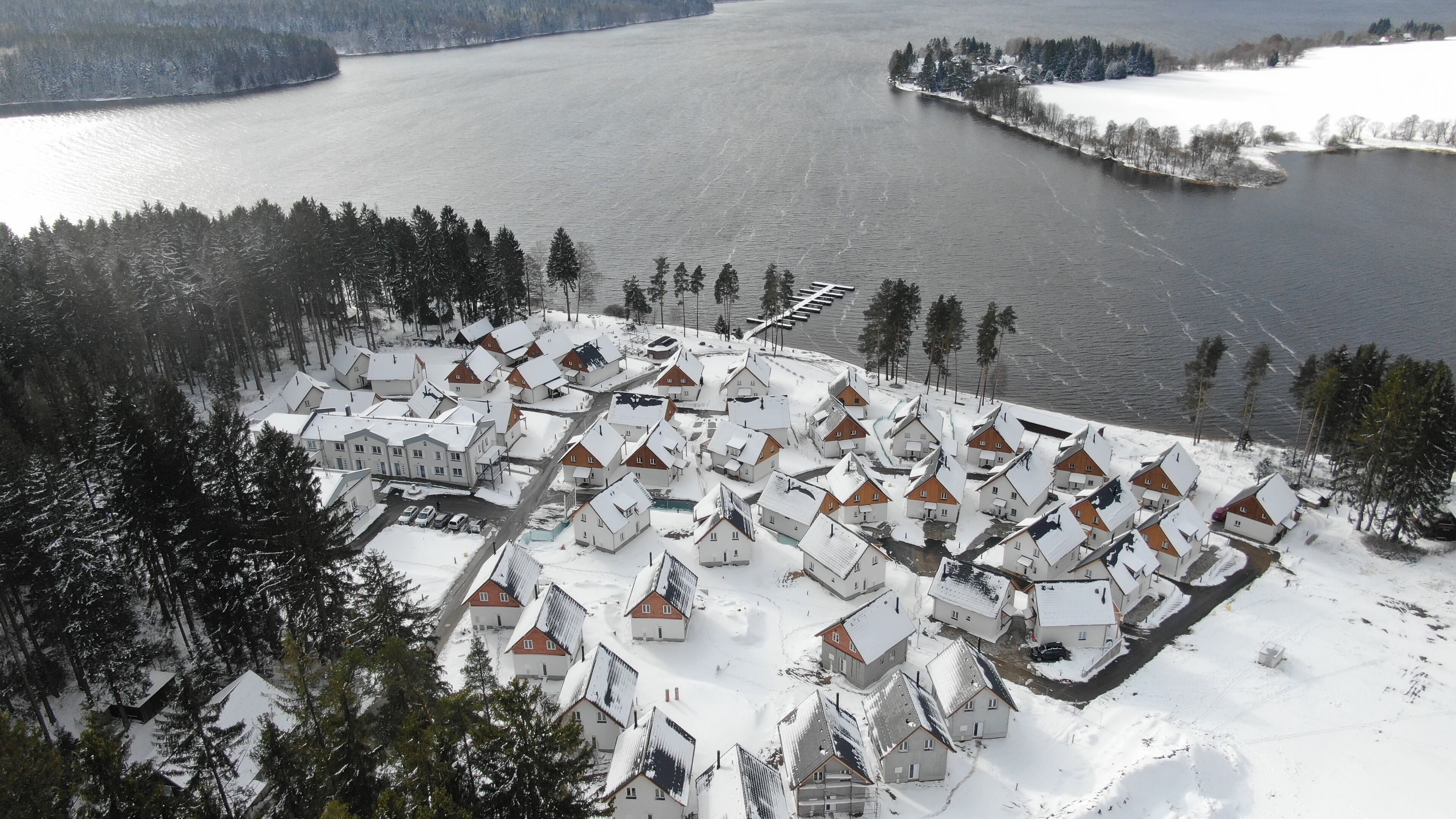 Ein schneebedecktes Dorf mit haussähnlichen Gebäuden in rasterförmiger Anordnung, ein zugefrorener See im Hintergrund und ein kleiner Steg, der sich in den See erstreckt.