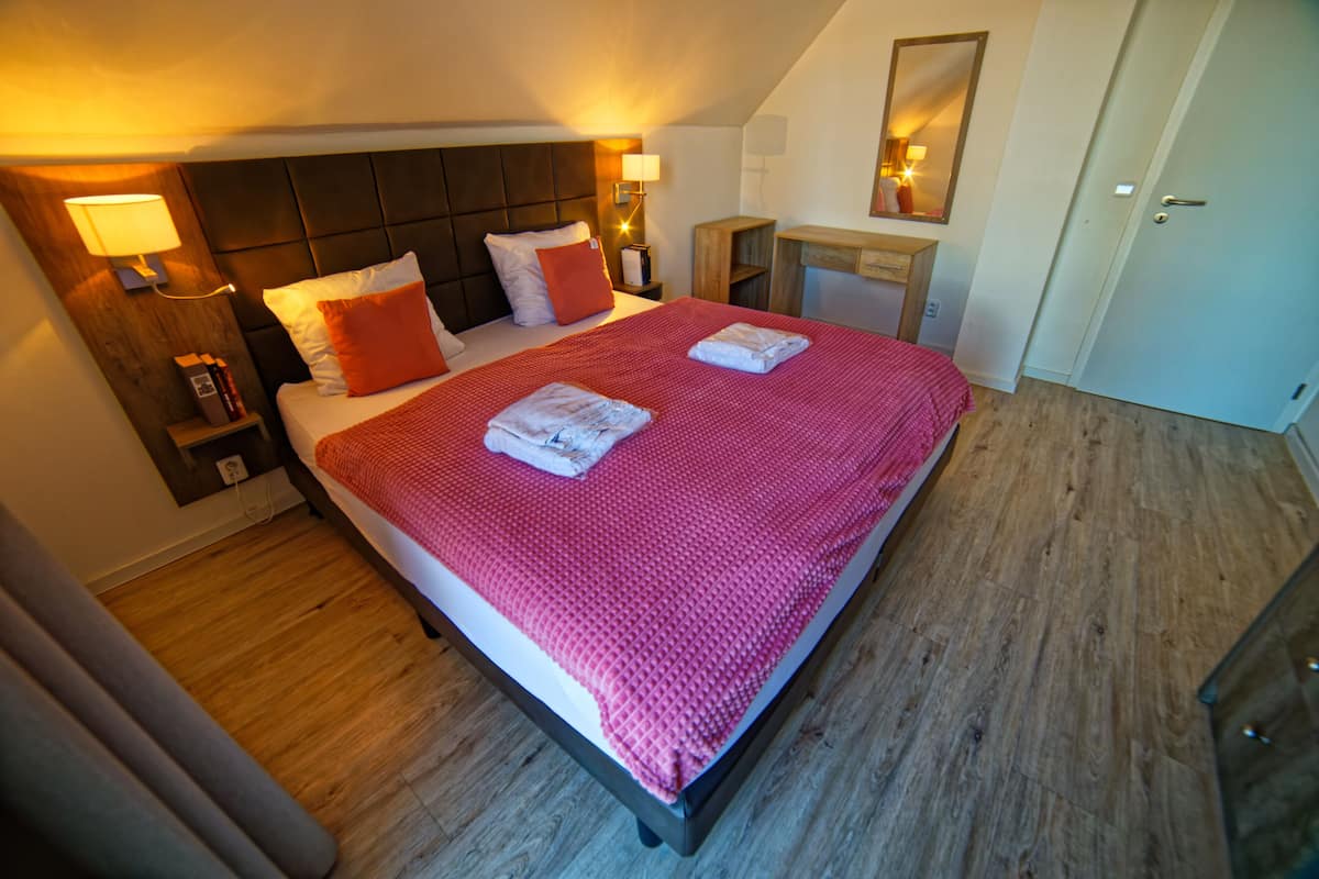 A neatly made bed with a red blanket and pillows, a bedside table with a lamp, a wooden nightstand, and a small desk with a mirror.