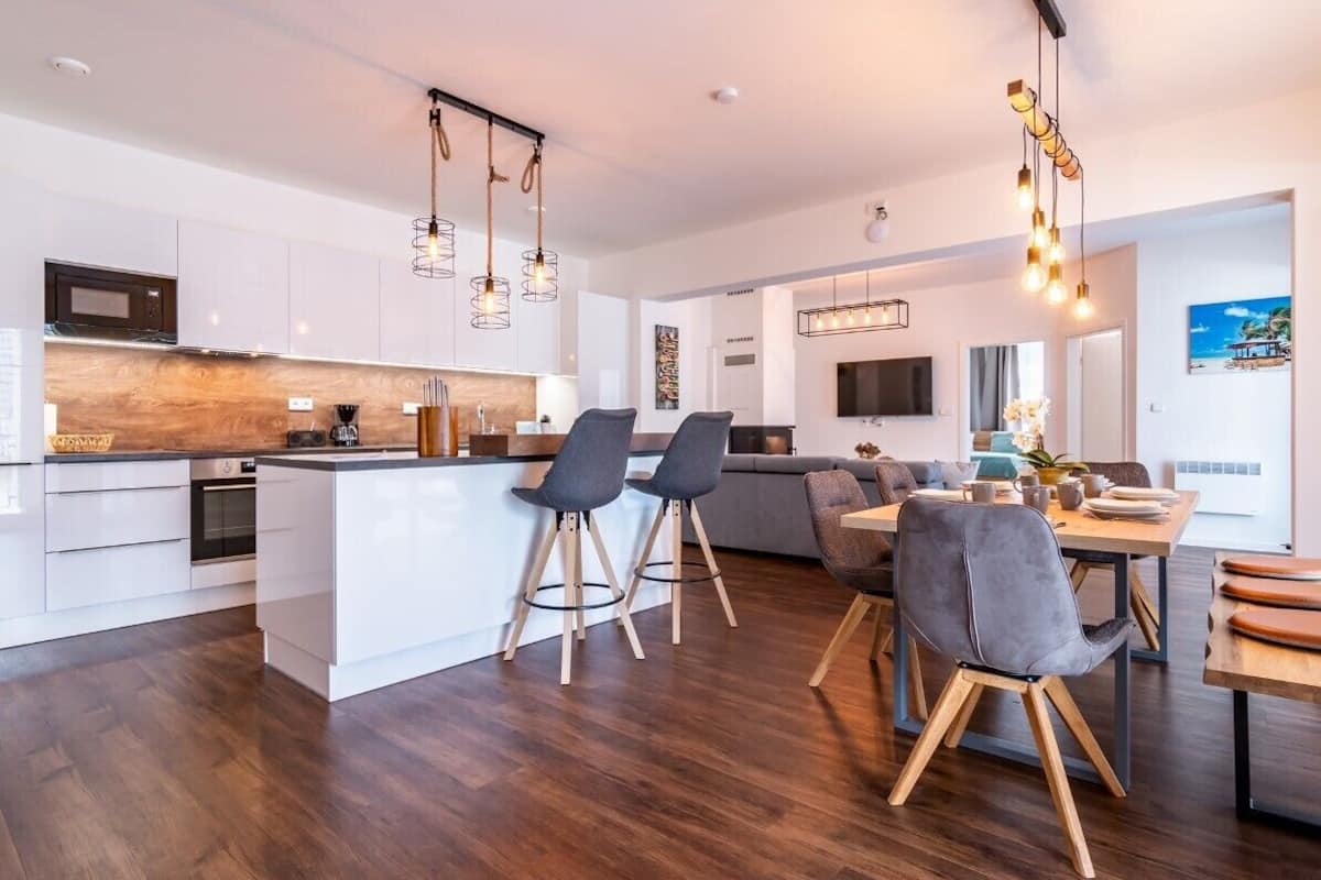 A modern kitchen with a central island, bar stools, a dining table with chairs, and a TV mounted on the wall.