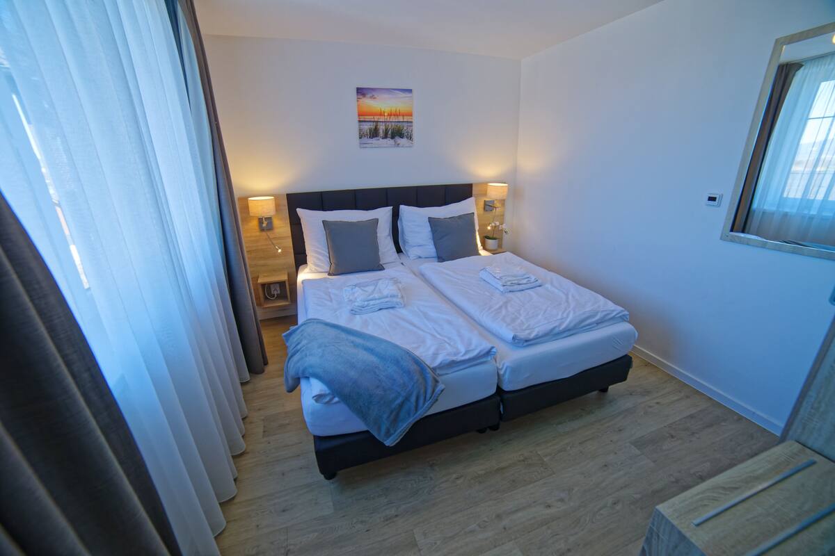 A double bed with two pillows, a bedside table with a lamp, and a window with curtains.