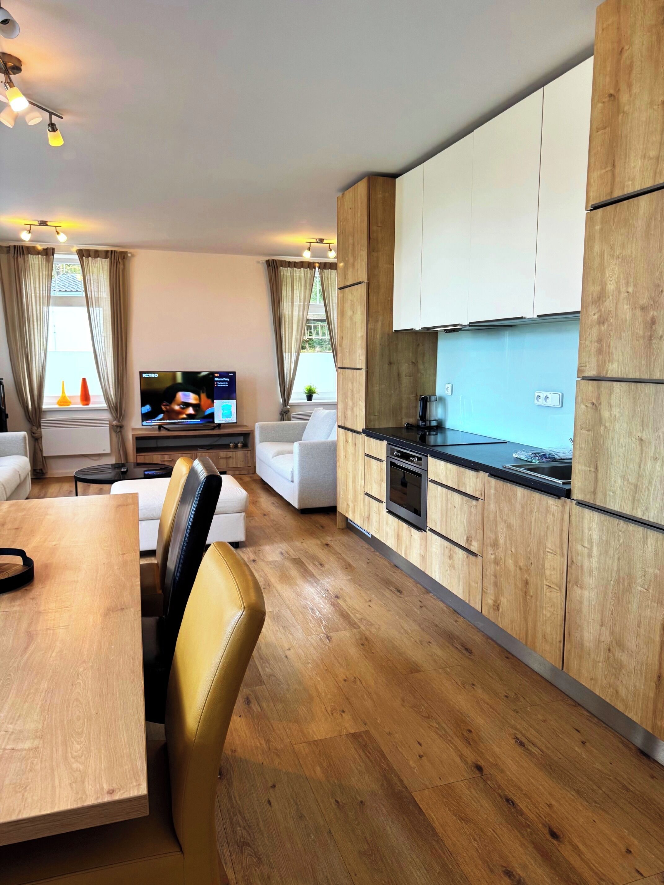 Eine moderne Küche mit Holzschränken und einer schwarzen Arbeitsplatte, ein Essbereich mit Holztisch und Stühlen sowie ein Wohnbereich mit Sofa und Fernseher.