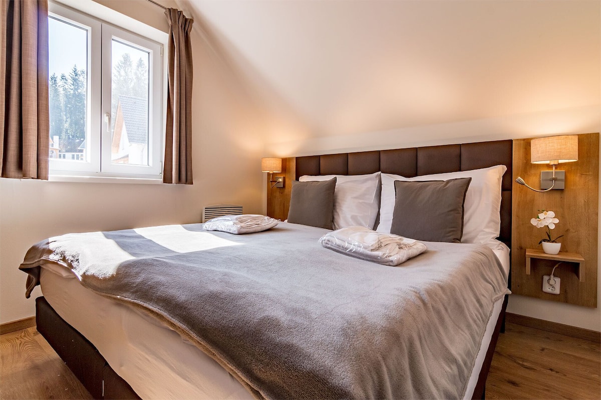 A neatly made bed with a grey blanket and pillows, a wooden headboard, bedside lamps, and a window with curtains.