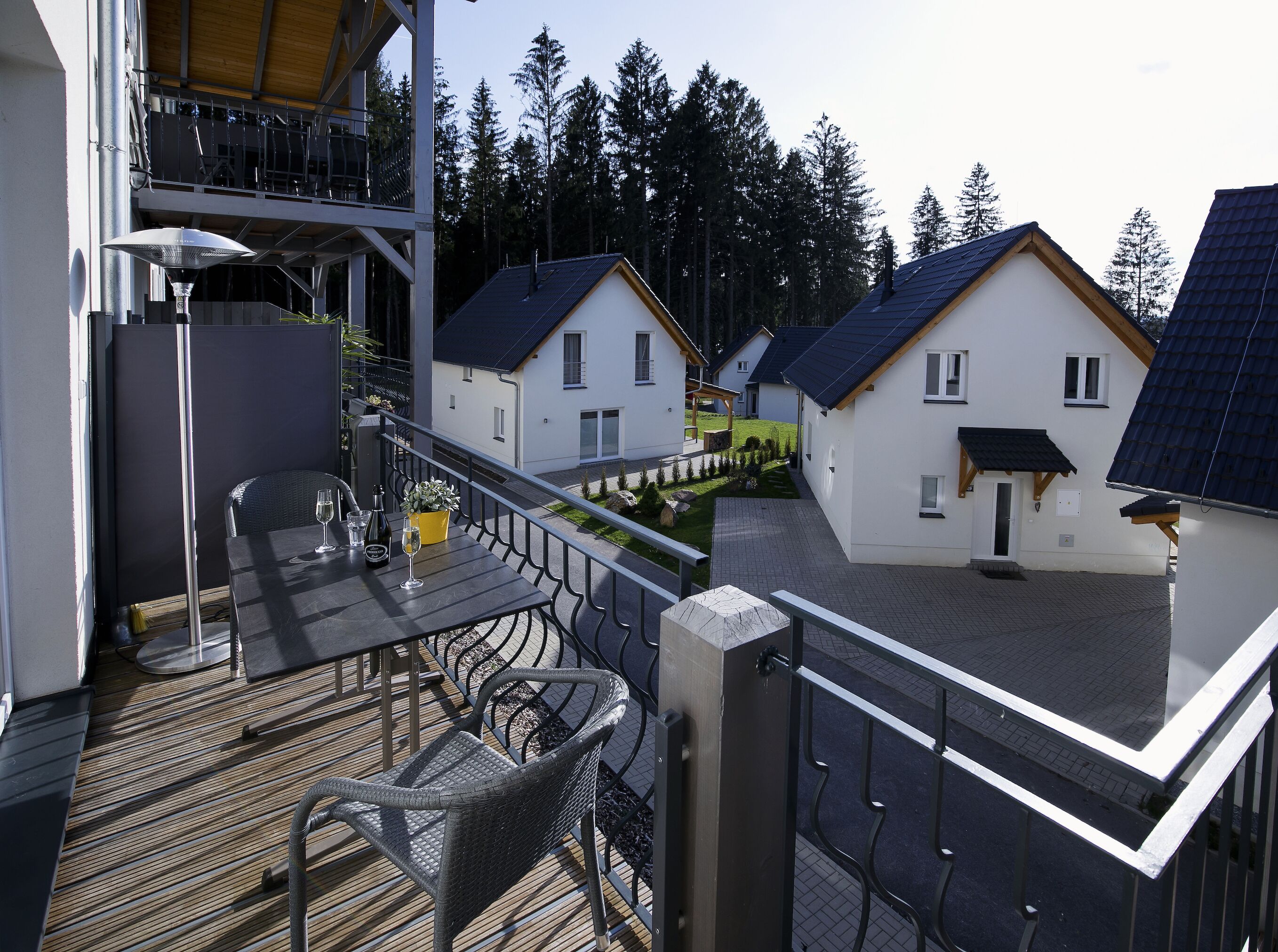 A balcony with a table and chairs, overlooking a row of houses with a forest in the background.