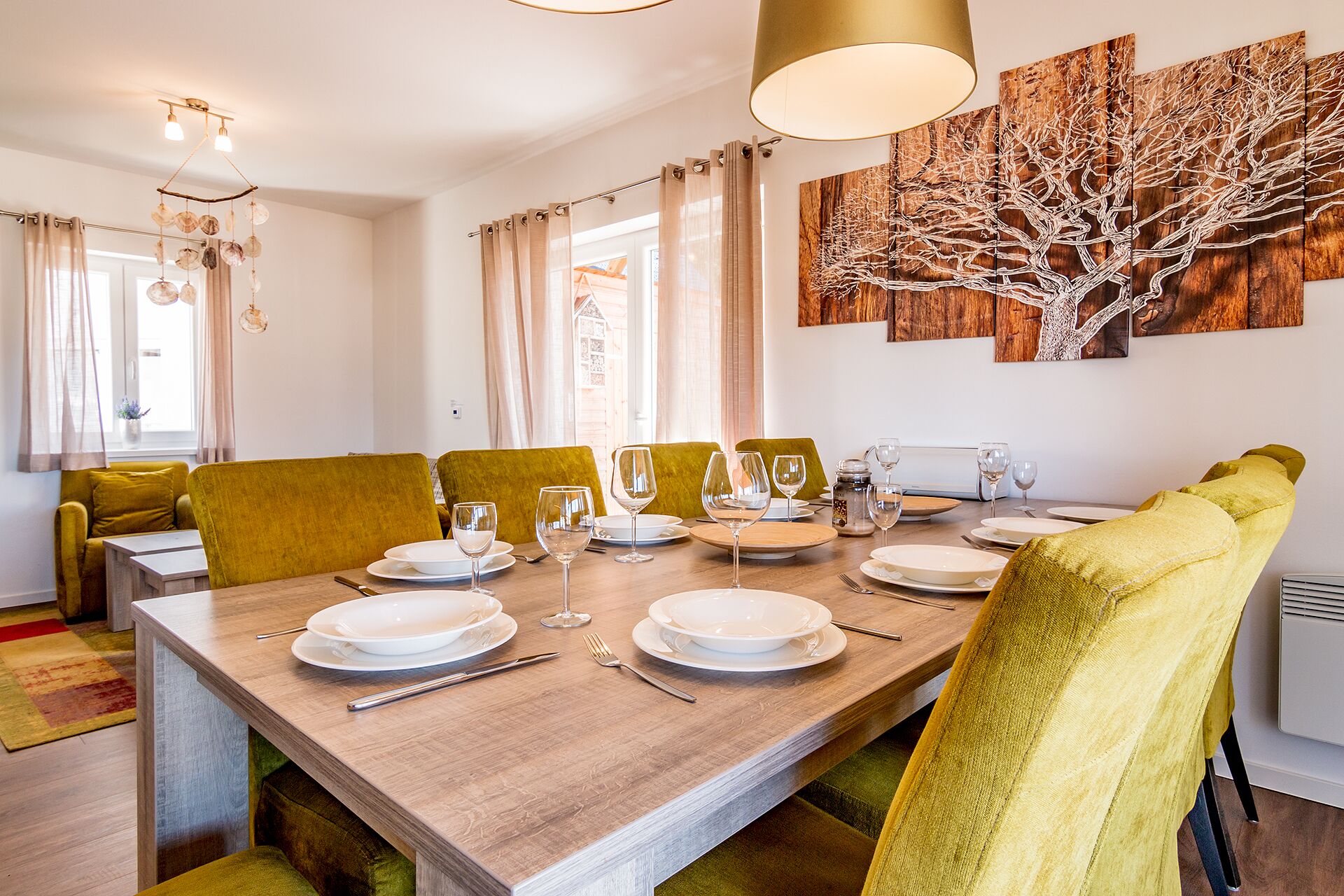 Un coin repas avec une longue table en bois, dressée pour quatre personnes, des chaises à l’assise rembourrée de tissu vert et un grand tableau représentant un arbre abstrait accroché au mur.