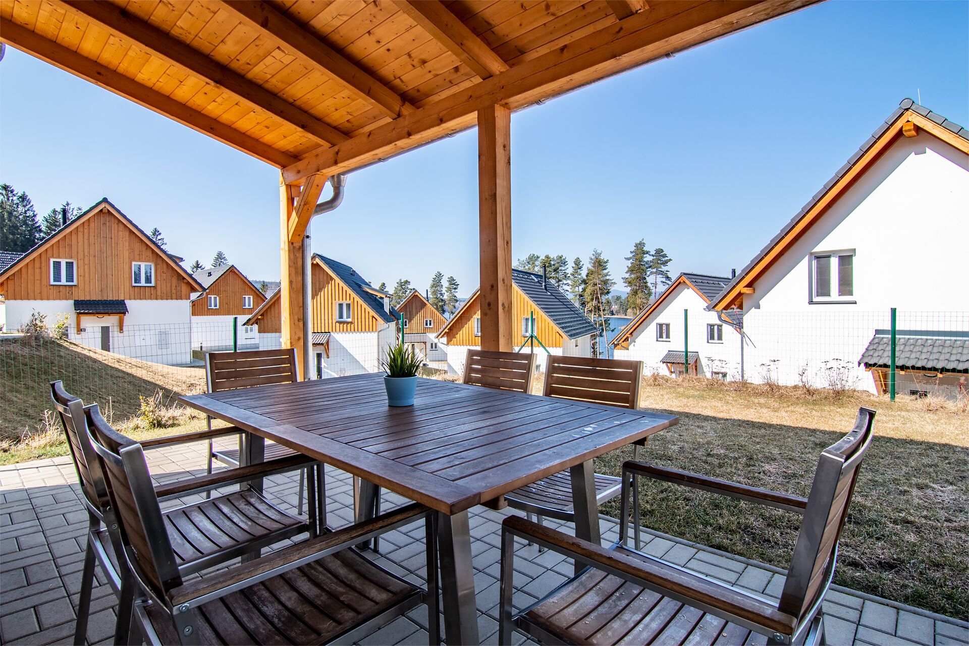 A covered outdoor area with a wooden table and benches, surrounded by houses with sloped roofs.