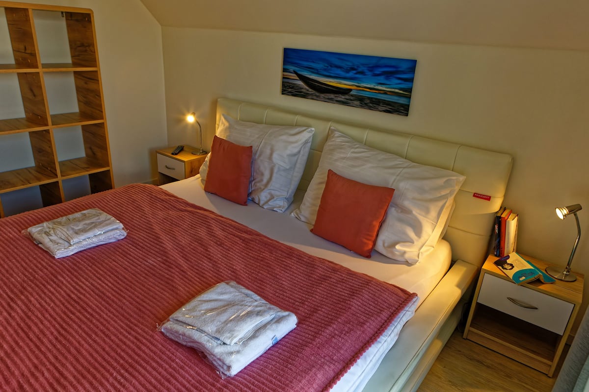 A neatly made bed with a red blanket, two pillows, and two folded towels. There is a nightstand with a lamp and a book. A painting of a boat on water is on the wall.