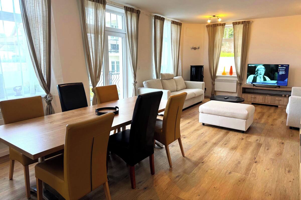 A modern living room with a wooden dining table, chairs, a sofa, and a television.
