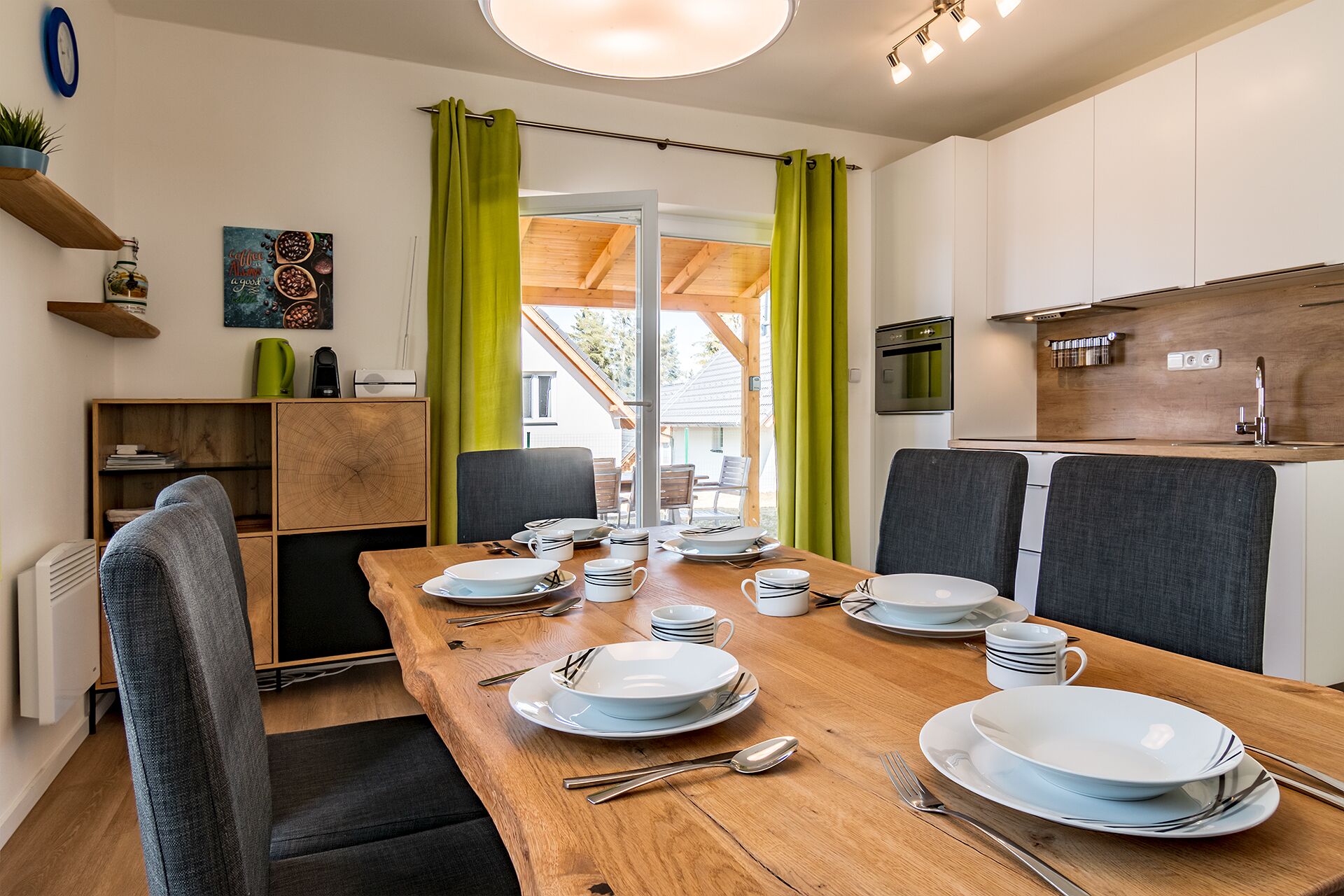 A dining area with a wooden table set for four, surrounded by grey upholstered chairs. A kitchen with white cabinets and a stainless steel sink is visible in the background.