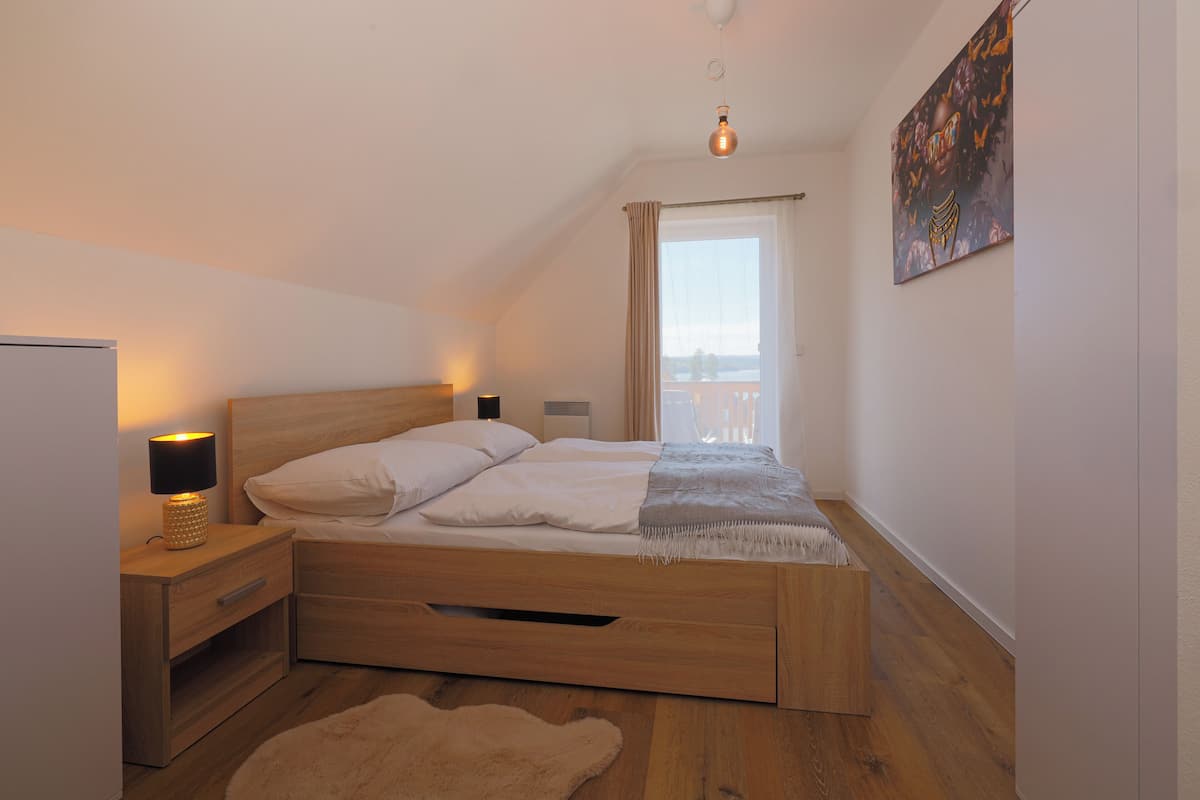 A bedroom with a wooden bed, a nightstand with a lamp, a window with curtains, and a painting on the wall.