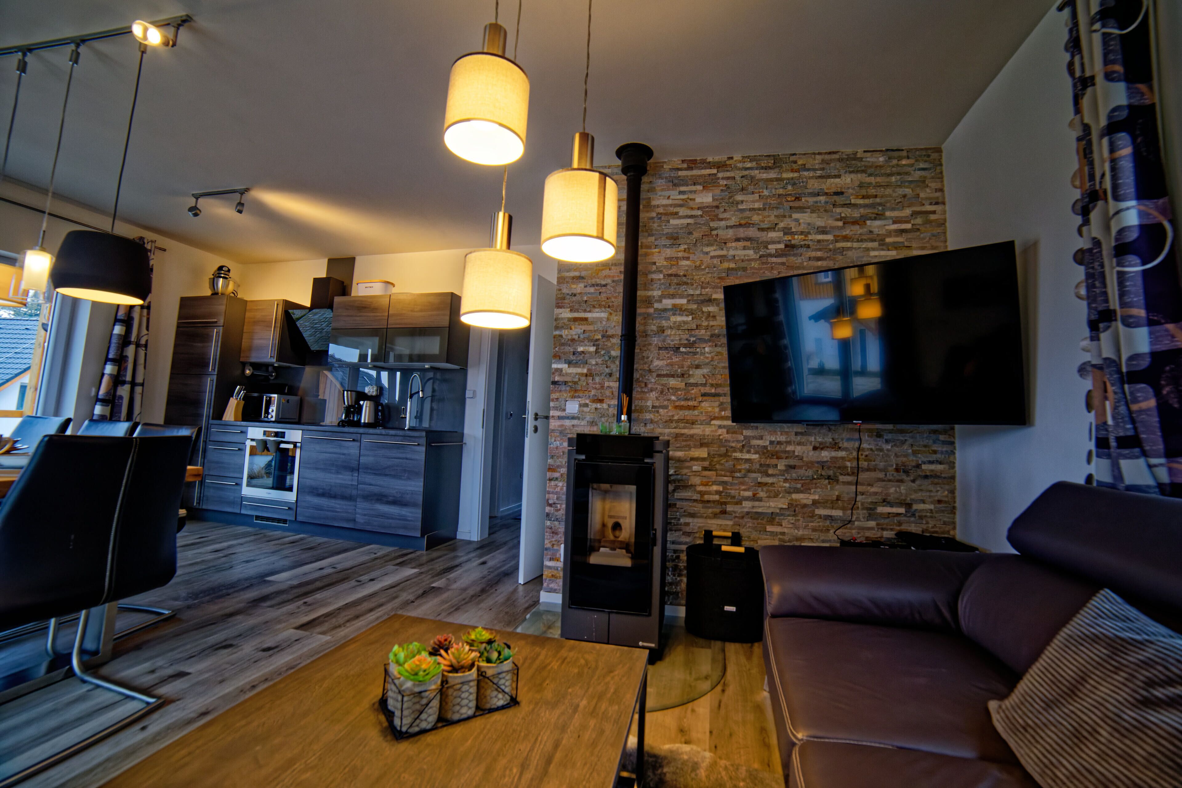 Un salon moderne doté d’une cheminée, d’un téléviseur à écran plat fixé à un mur en briques, et d’un coin repas avec une table et des chaises en bois.