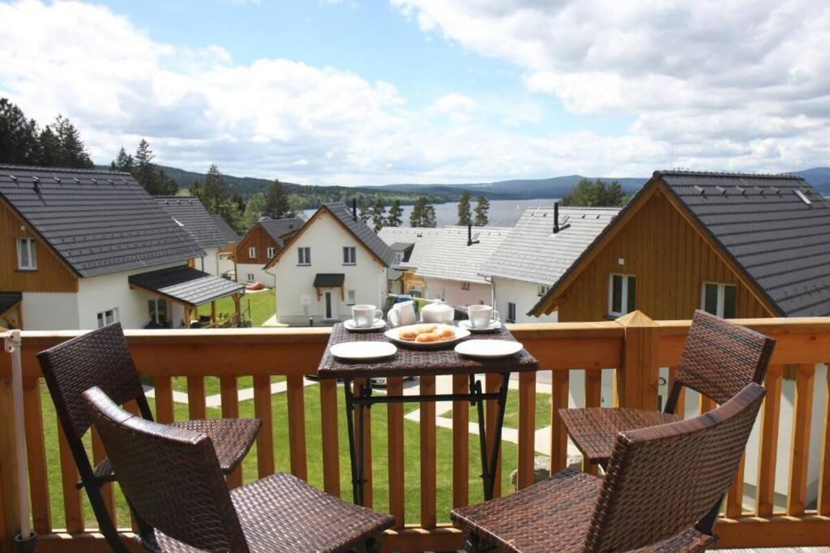 A balcony with wicker chairs and a table set for breakfast, overlooking a row of houses and a lake.