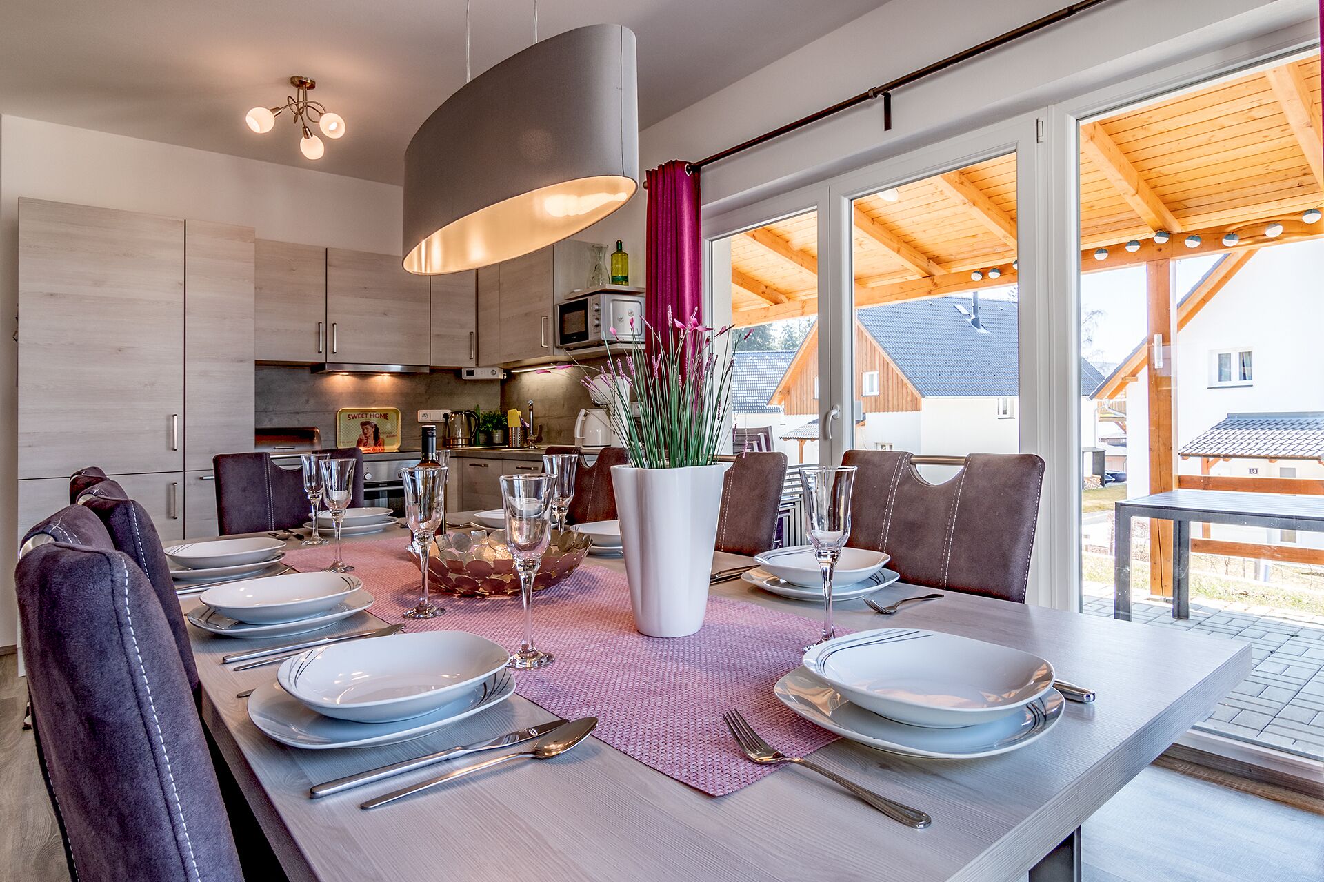 Un comedor moderno con una larga mesa dispuesta para seis comensales, un jarrón central con flores y grandes ventanales que ofrecen vistas del entorno.