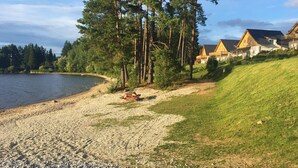 A lakeside property with a gravel path leading to a wooden deck, surrounded by tall pine trees and a row of wooden houses with sloped roofs. - Lakeside Village Větrník (Frymburk)