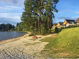 Una propiedad frente al lago con un camino de grava que lleva a una terraza de madera, rodeada de altos pinos y una hilera de casas de madera con techos inclinados.