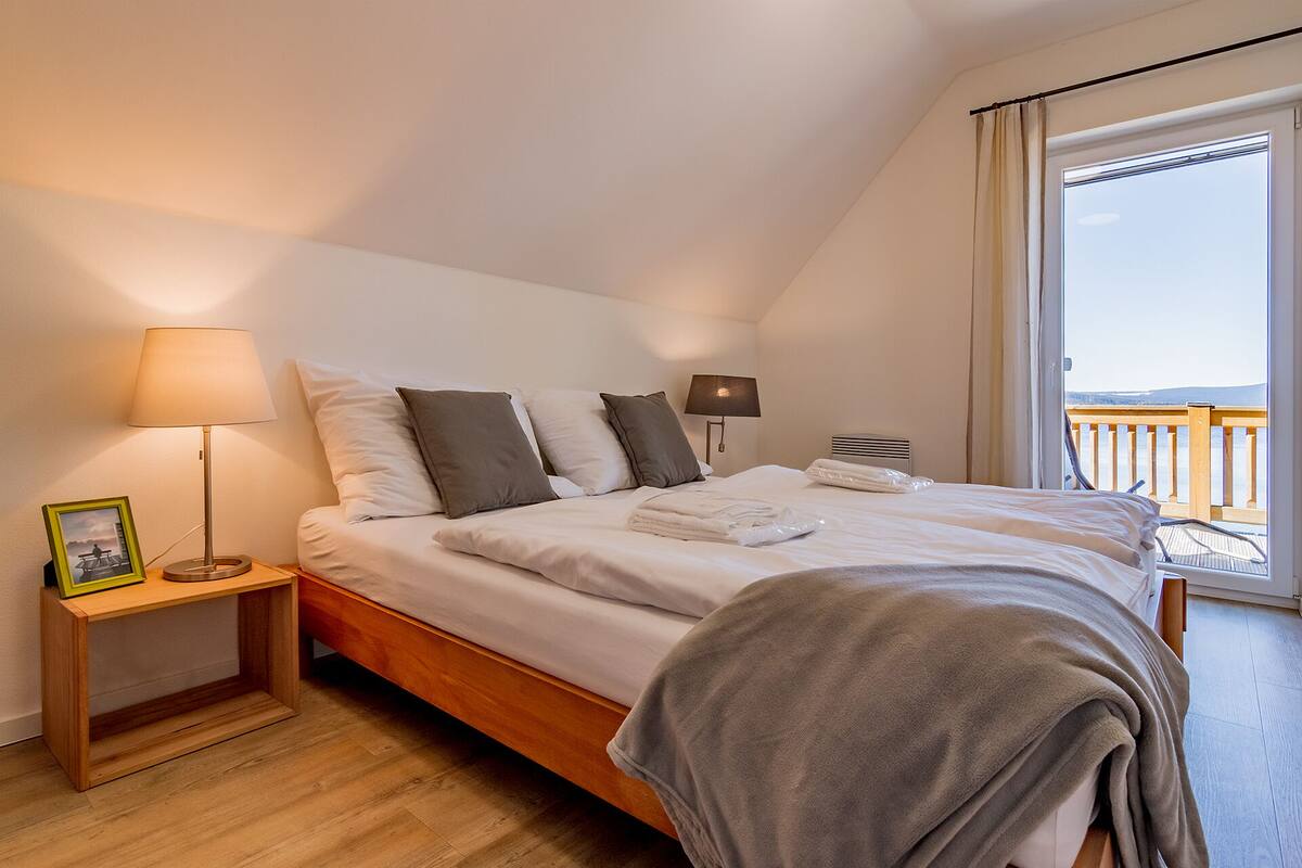 A bedroom with a bed, two bedside lamps, a wooden nightstand, a framed picture, and a view of the sea through a balcony.