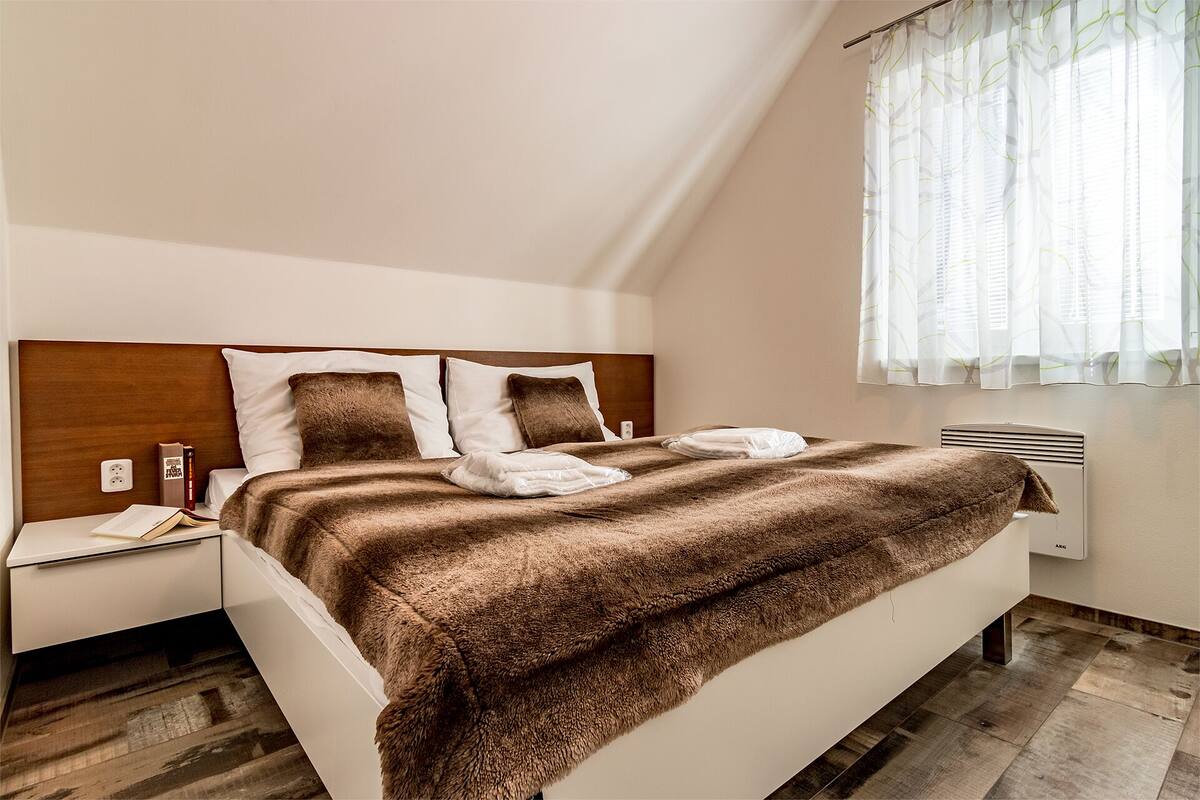A neatly made bed with a brown blanket and pillows, a bedside table with a book and a power outlet, and a window with sheer curtains.
