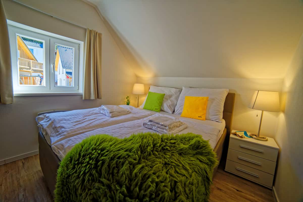 A bedroom with a bed, two bedside lamps, a nightstand, a window with curtains, and a green furry ottoman.