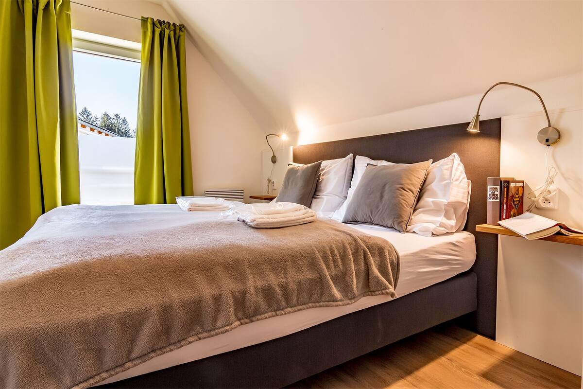 A neatly made bed with a grey blanket and pillows, a bedside table with books and a lamp, and a window with green curtains.