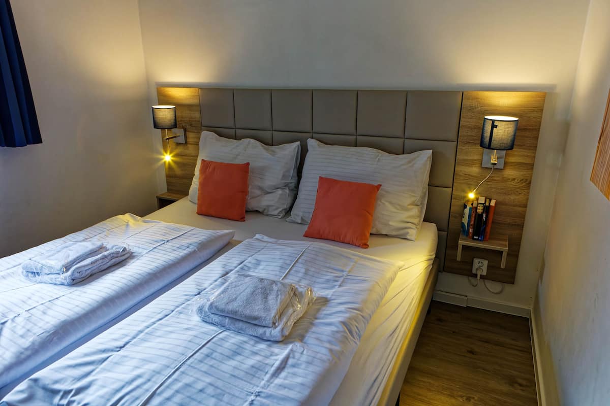 A hotel room with two beds, each with a pillow and a folded towel. There are wall-mounted lamps on either side of the beds.