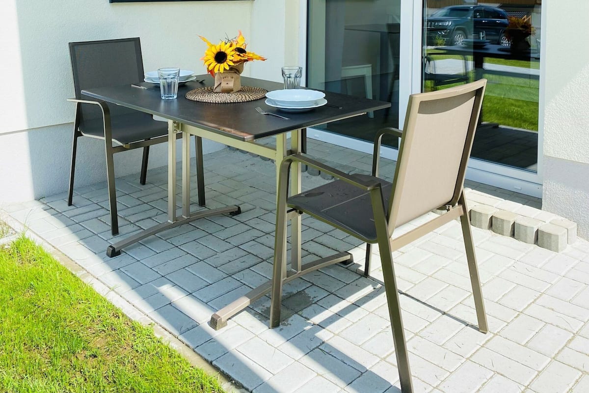 A patio with a table and chairs, a window with plants, and a view of houses through the glass.