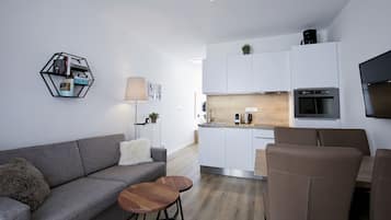 Una sala de estar moderna con un sofá gris, una mesa de centro de madera y una estantería hexagonal. En el fondo, se ve una cocina con armarios blancos, encimera de madera y un horno empotrado.
