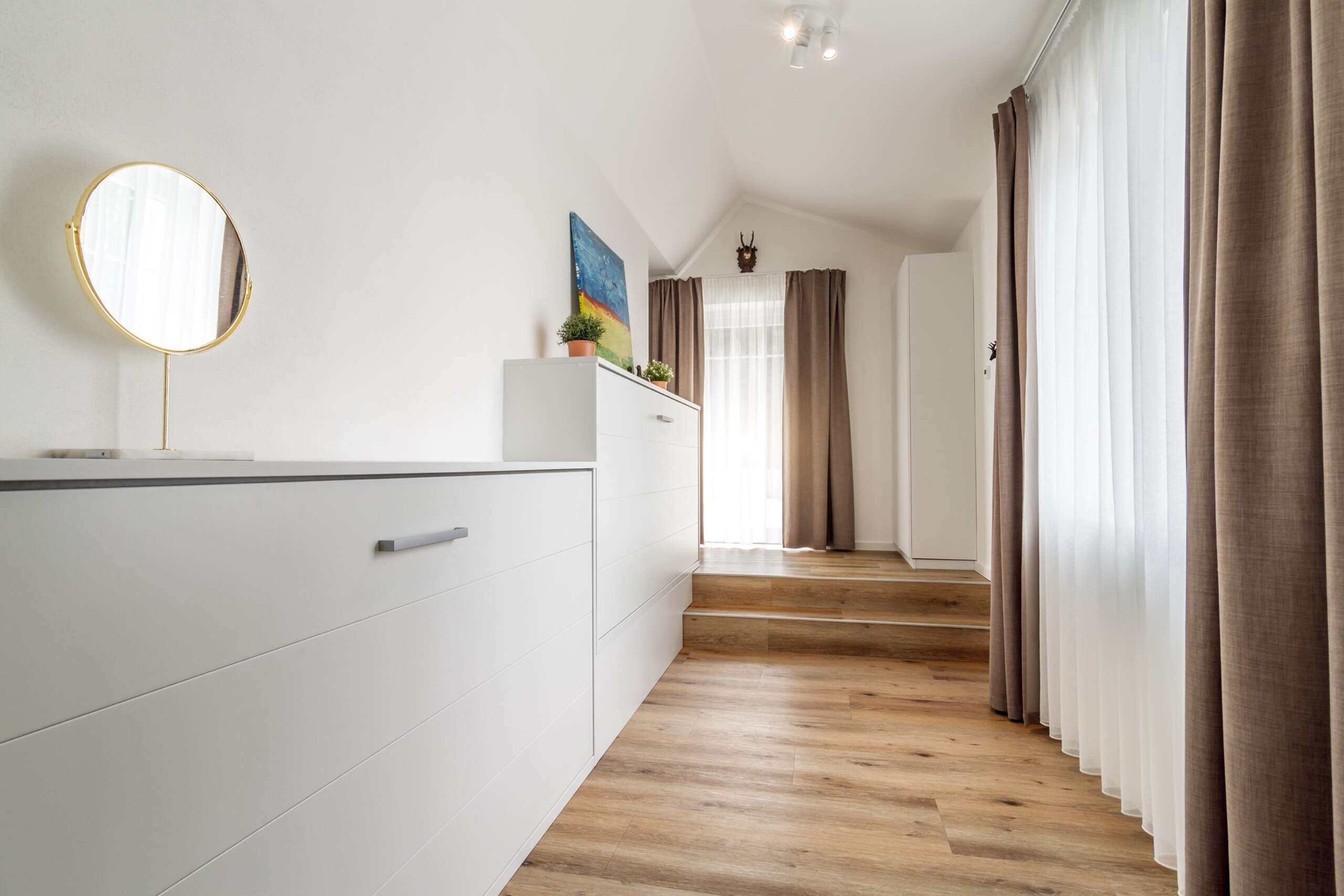 Una habitación de hotel moderna con suelo de madera, paredes blancas, un espejo grande, una planta pequeña y una escalera que lleva a otra habitación.