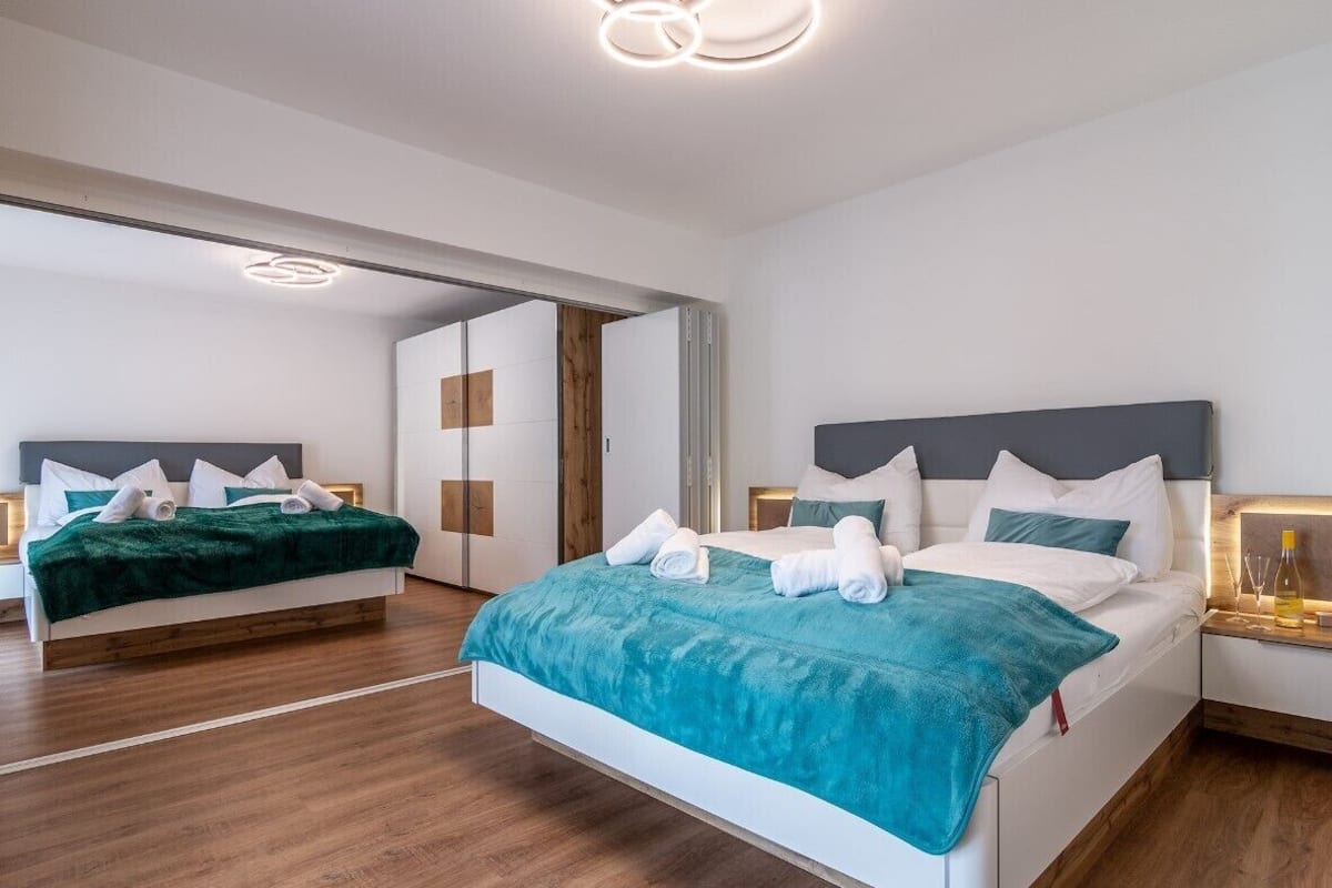 A modern hotel room with two beds, wooden flooring, and a bright ceiling light fixture.