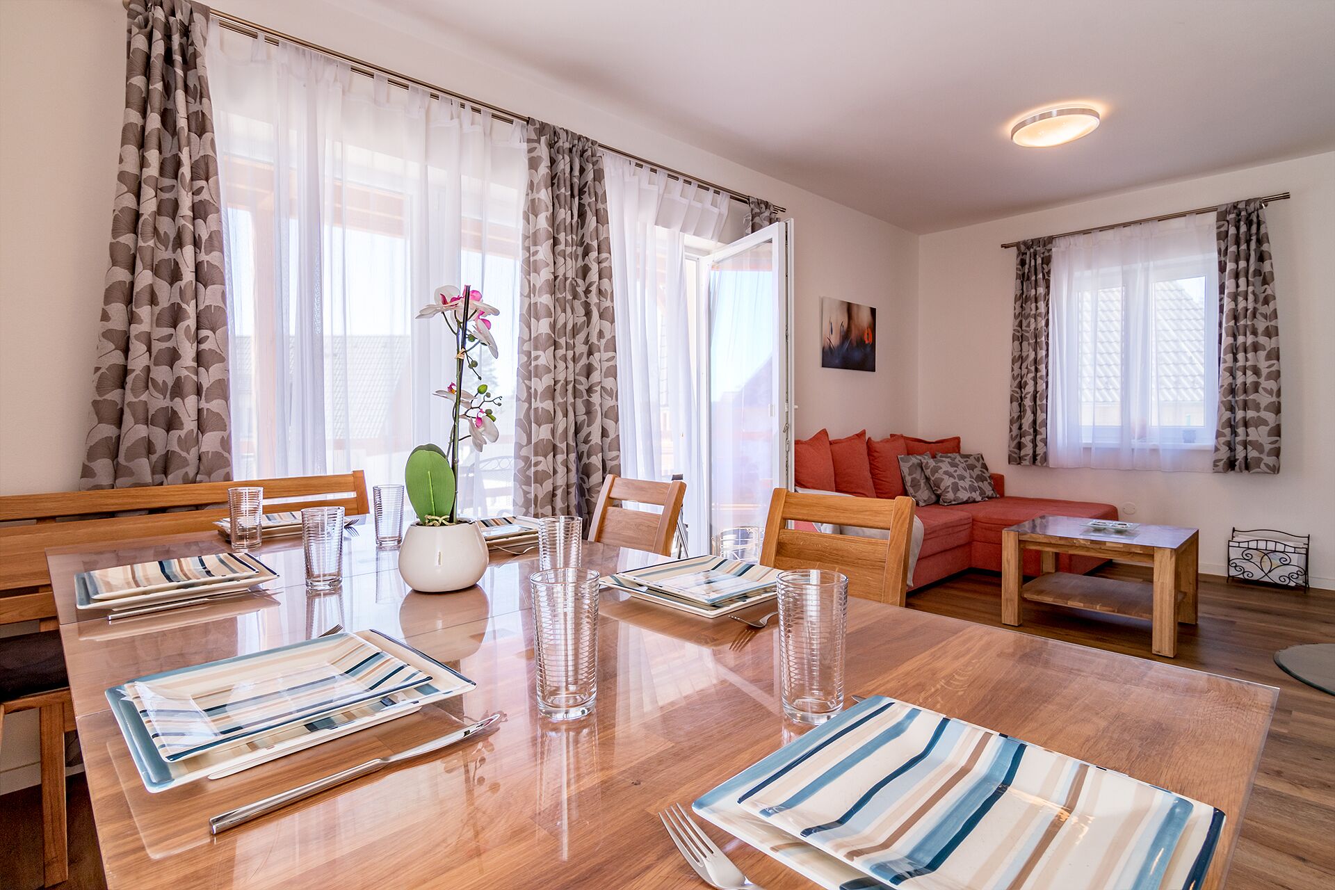 Un comedor con una mesa de madera, sillas y un jarrón con flores. Se observa un sofá y una mesita en el fondo.