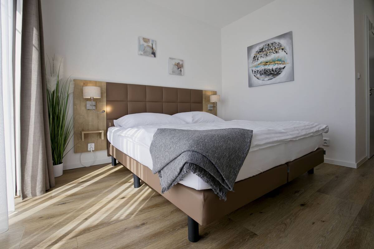 A modern bedroom with a large bed, wooden floor, and a headboard with wall-mounted lights.