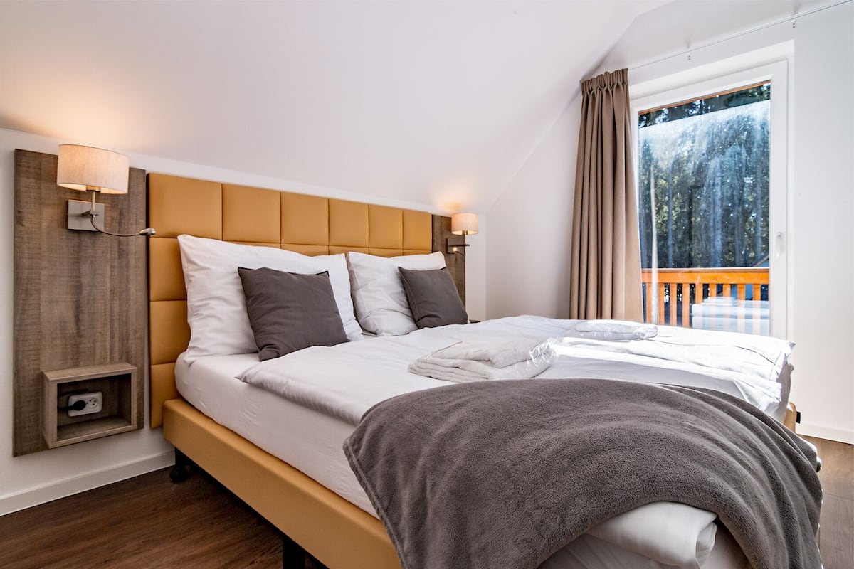A modern bedroom with a wooden headboard, a bedside table with a lamp, and a window with curtains.
