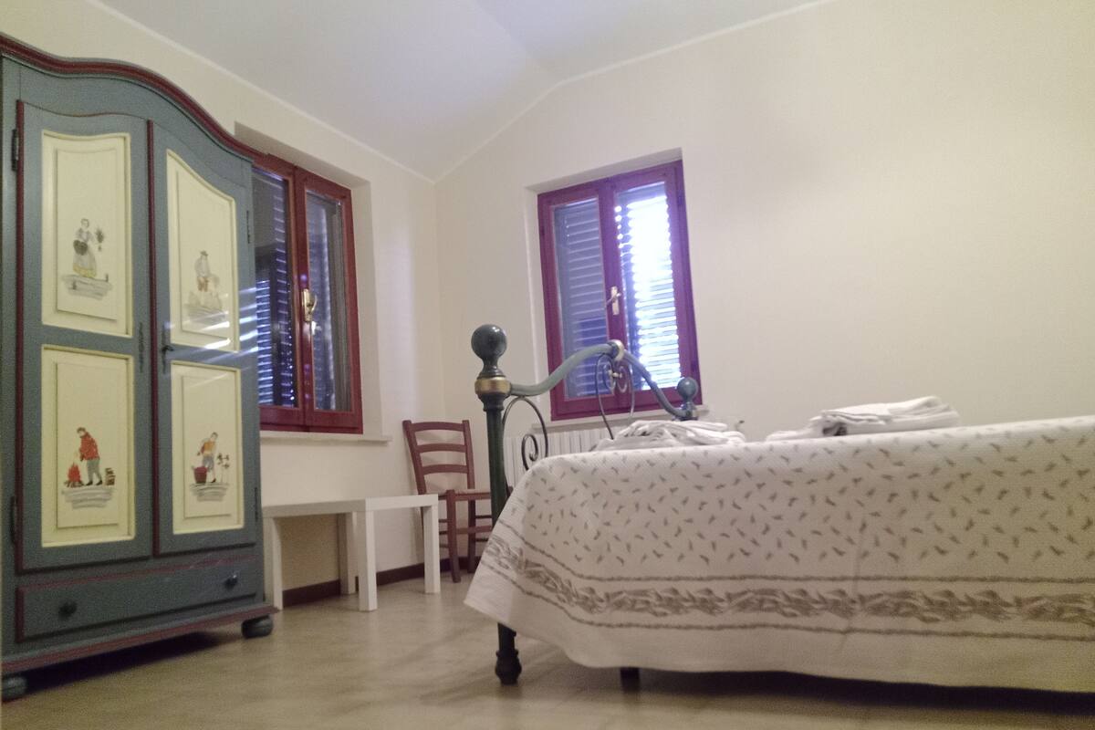Ein Schlafzimmer mit einem Bett, einem Schminktisch mit Kunstwerken, einem Stuhl und einem Fenster mit Jalousien.