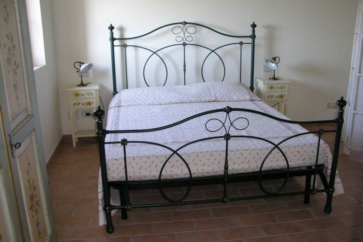 Ein Bett mit Metallgestell und einem gemusterten Bettbezug in einem Zimmer mit Fliesenboden und weißen Wänden.