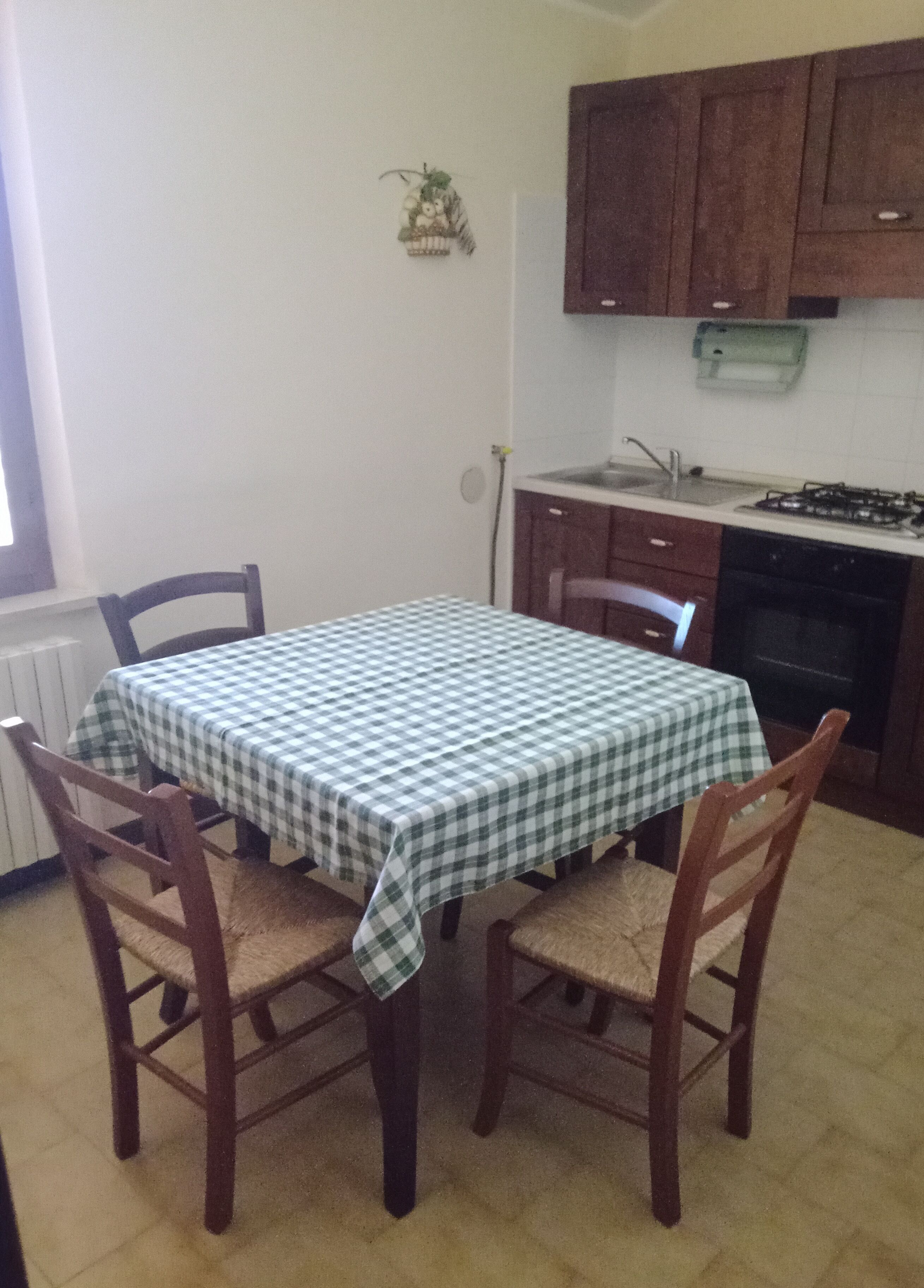 Una mesa de comedor con mantel a cuadros, rodeada de sillas de madera. Se ve una cocina con armarios de madera y una estufa al fondo.