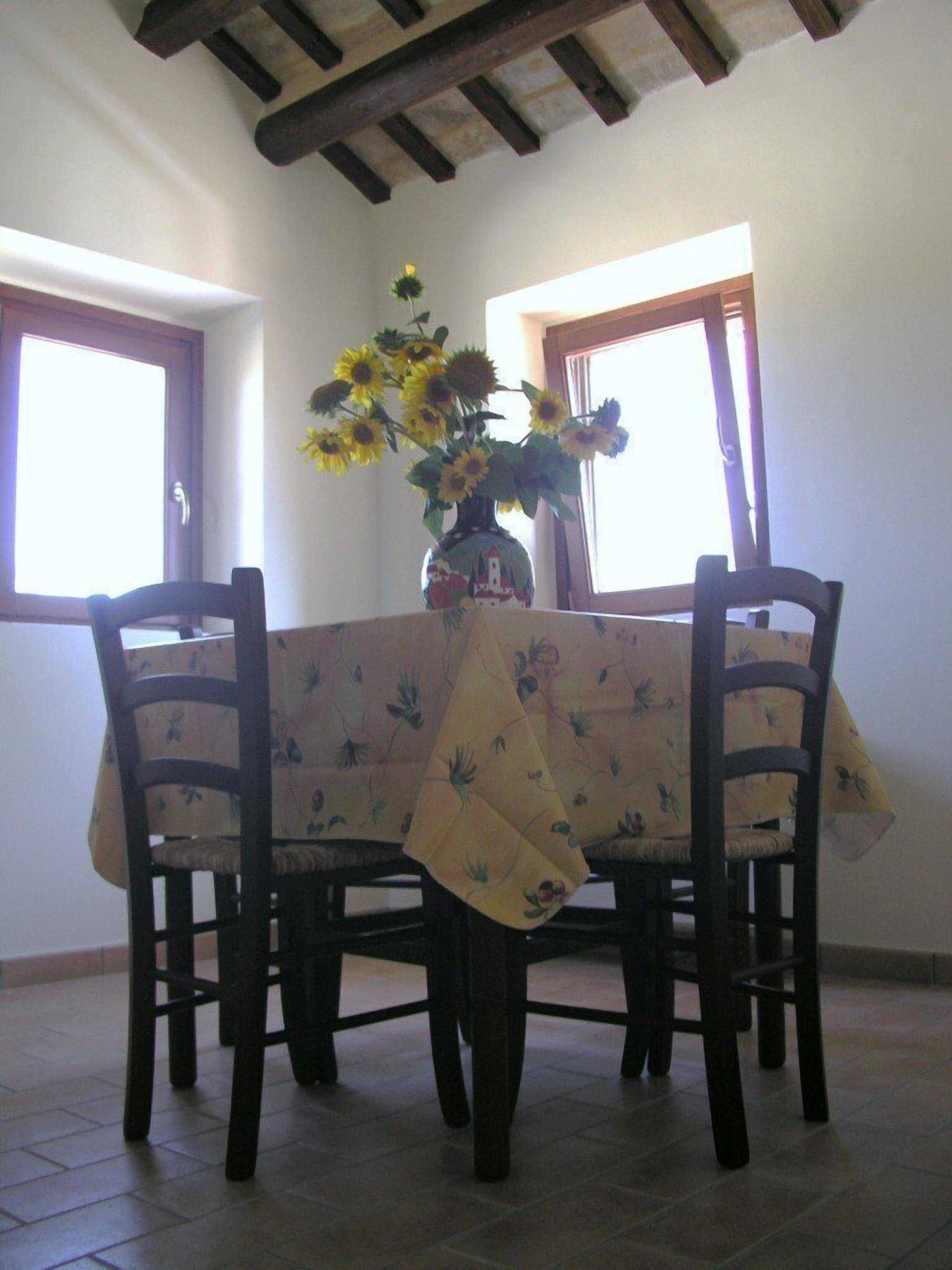 A dining table with a floral tablecloth, two chairs, and a vase with sunflowers.