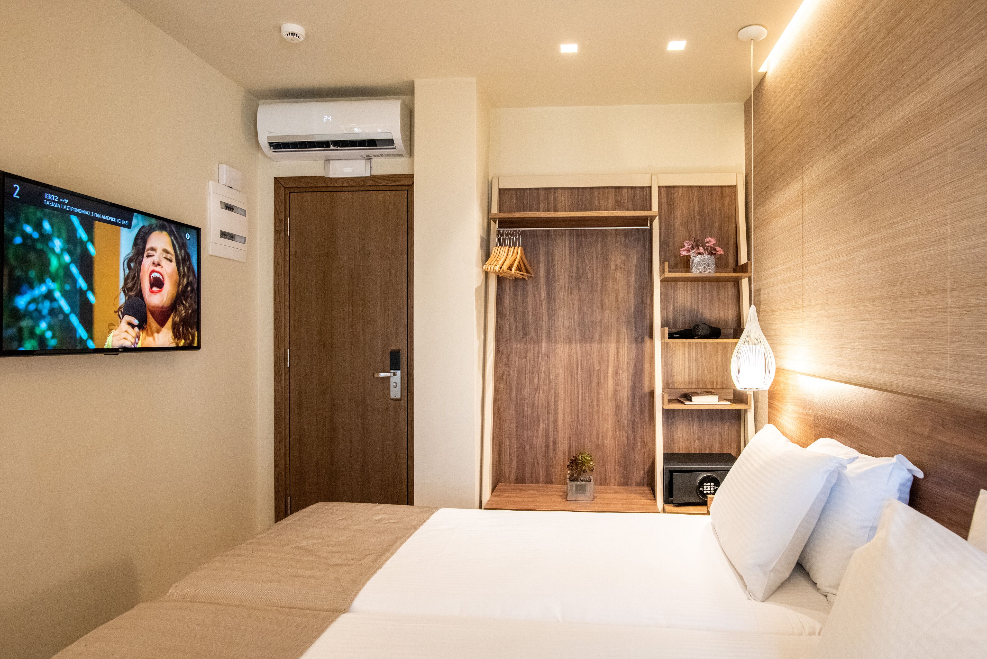 A hotel room with a bed, a TV mounted on the wall, a wooden wardrobe, and a bedside table with a lamp.