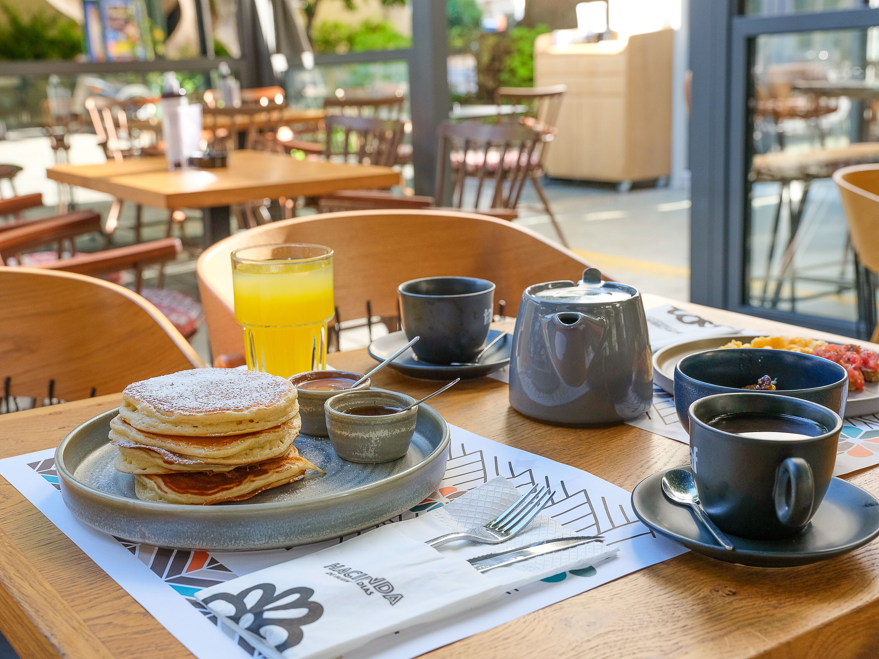 A breakfast setting with pancakes, a coffee pot, cups, and a glass of orange juice on a table.