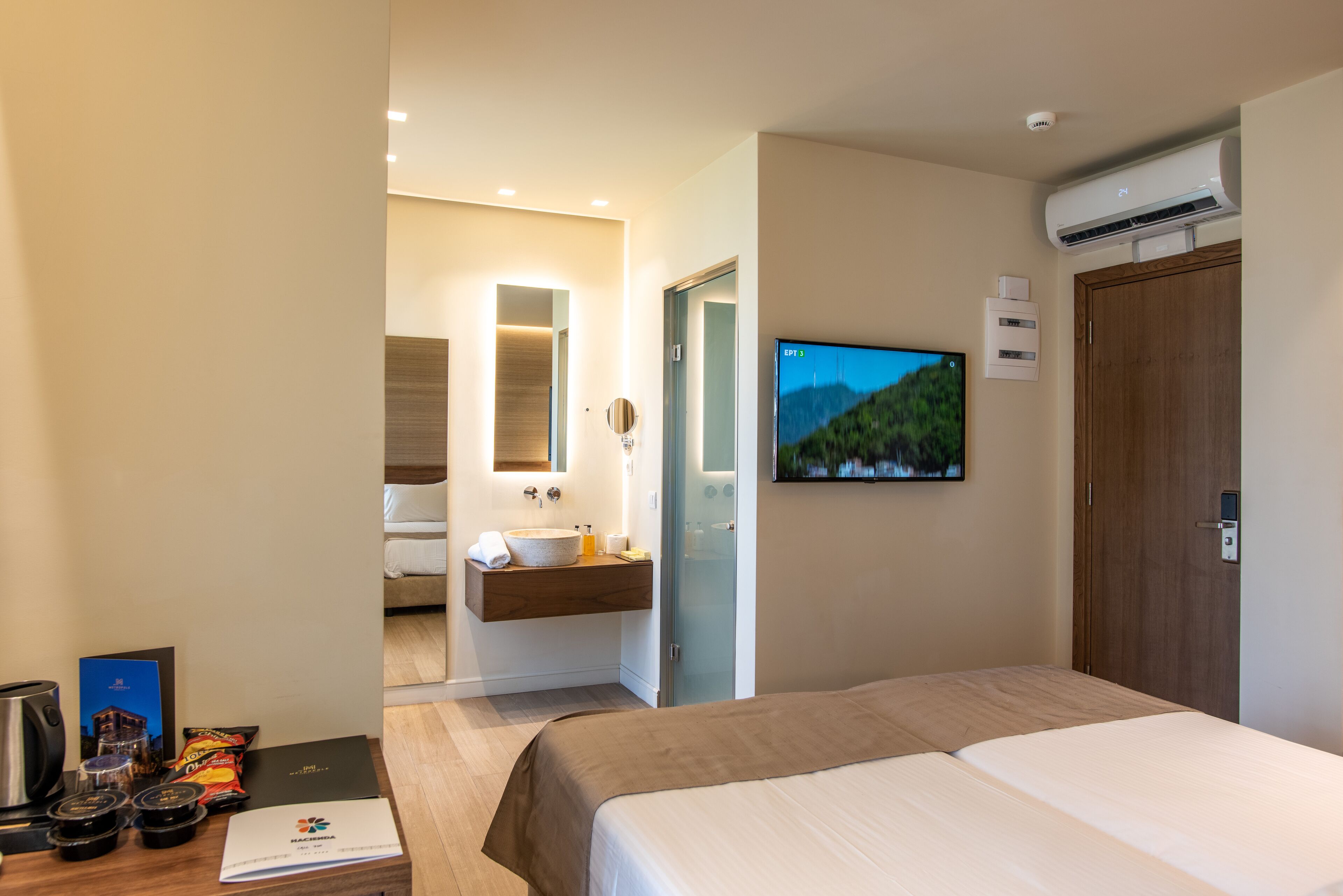A modern hotel room with a large bed, a bathroom with a sink and mirror, a television mounted on the wall, and an air conditioning unit.