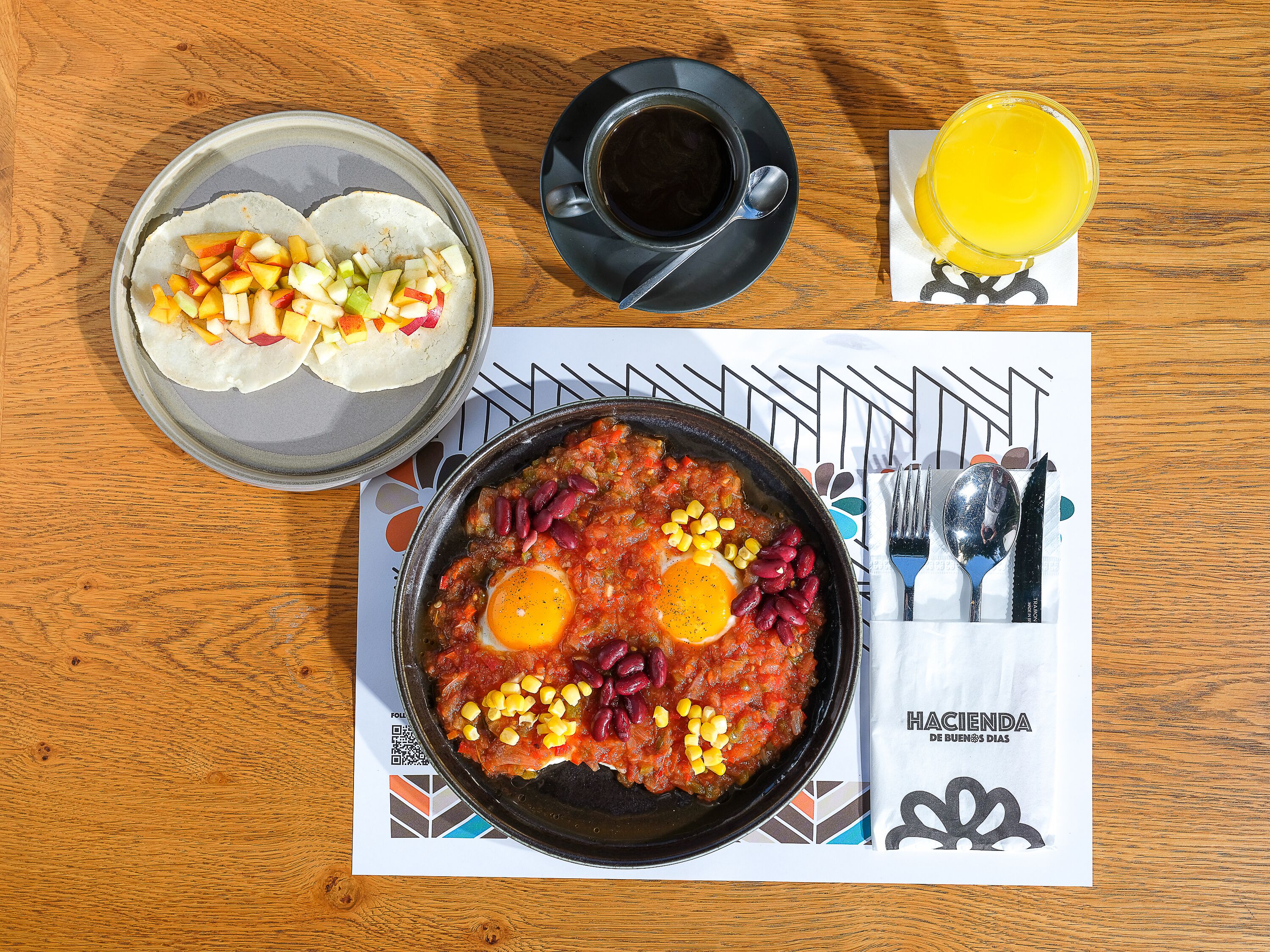 A meal with a bowl of beans and corn, a plate of fruit, a cup of coffee, and a glass of juice on a wooden table.