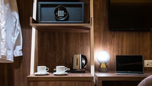 Una mesita de noche de madera con una cafetera, una taza, un control remoto y un ordenador portátil.