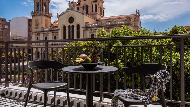 Un balcón con dos sillas, una mesa con frutero y vistas a un edificio histórico con torre del reloj y cúpula.