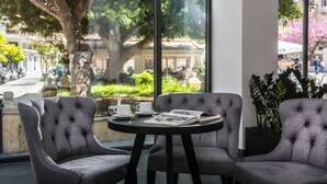 Un área de estar moderna en interior con una mesa redonda y dos sillas tapizadas, una ventana con vistas a una calle y zonas verdes, y una planta en maceta a la derecha.