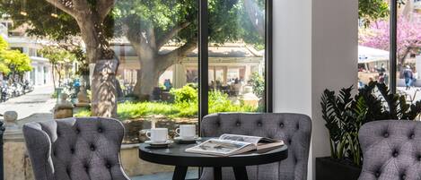 A modern indoor seating area with a round table and two upholstered chairs, a window with a view of a street and greenery, and a potted plant on the right.