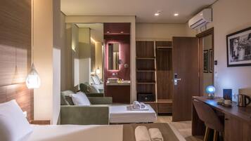 A hotel room with a bed, a desk, a chair, a sofa, a mirror, a wardrobe, and a bathroom visible through an open door.