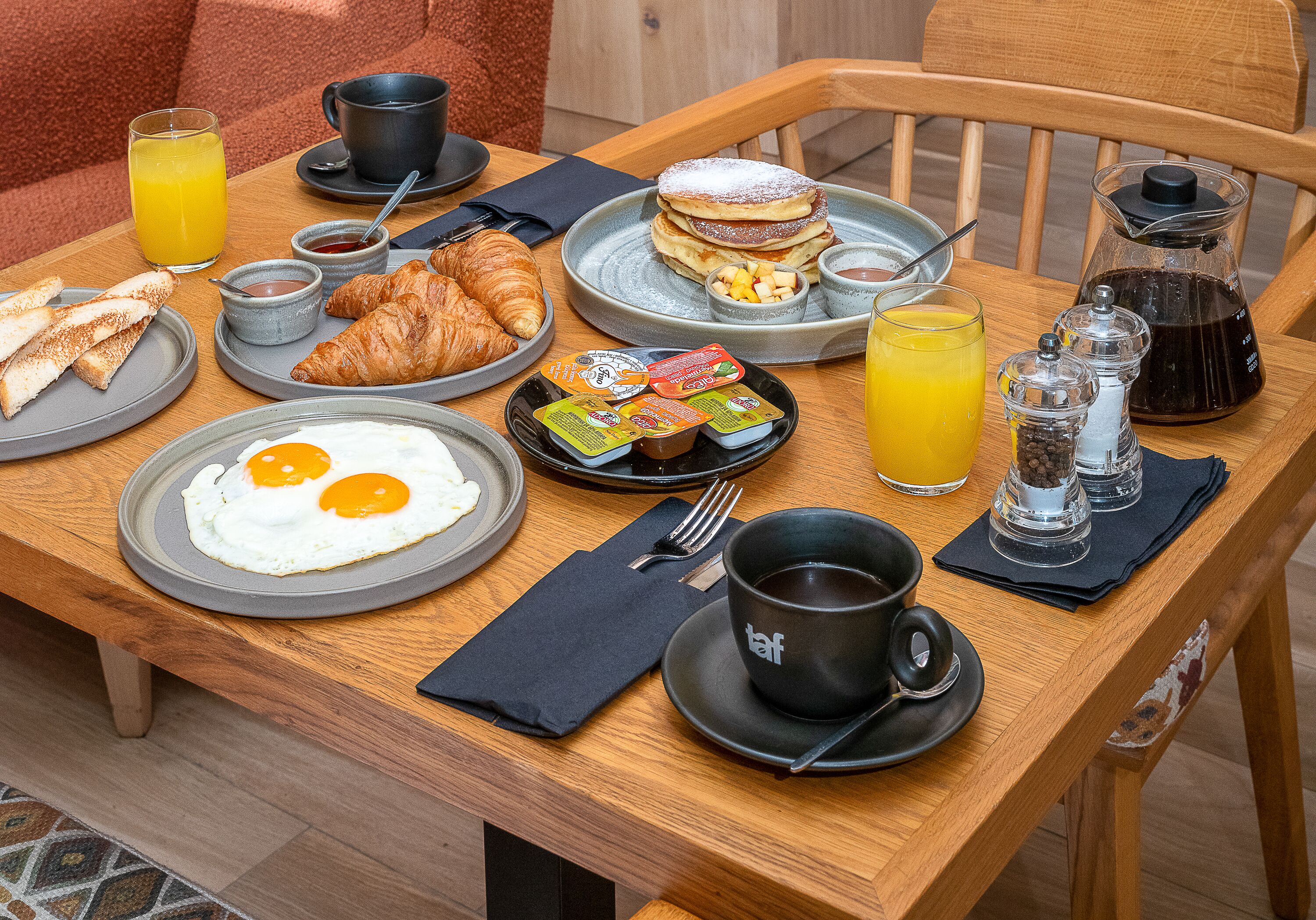 A breakfast table with plates of eggs, croissants, pancakes, and juice.