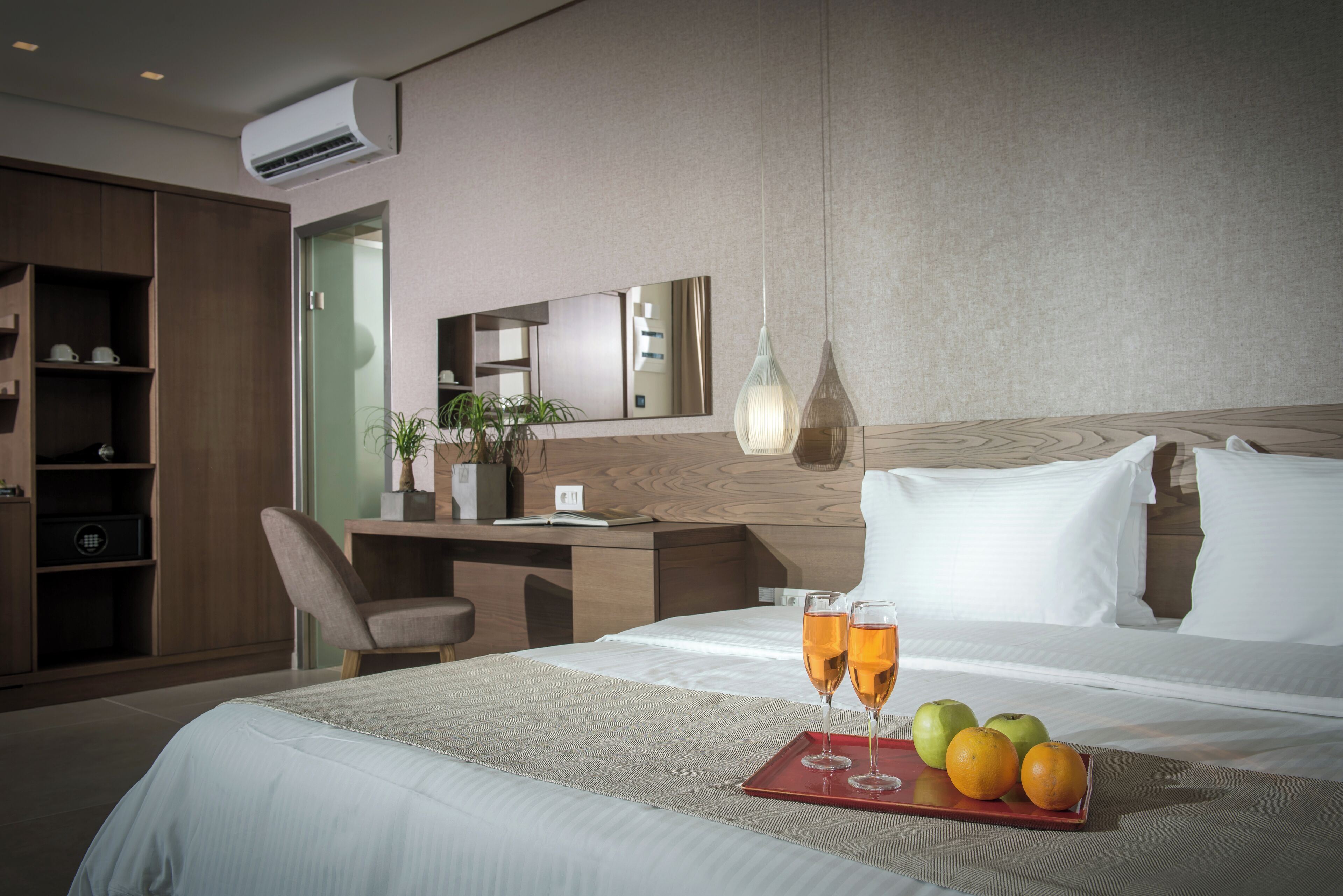 A hotel room with a bed, bedside table, and a desk with a chair. There is a tray with glasses and fruits on the bed.