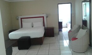 A hotel room with a bed, bedside tables, a small stool, and a wicker chair.